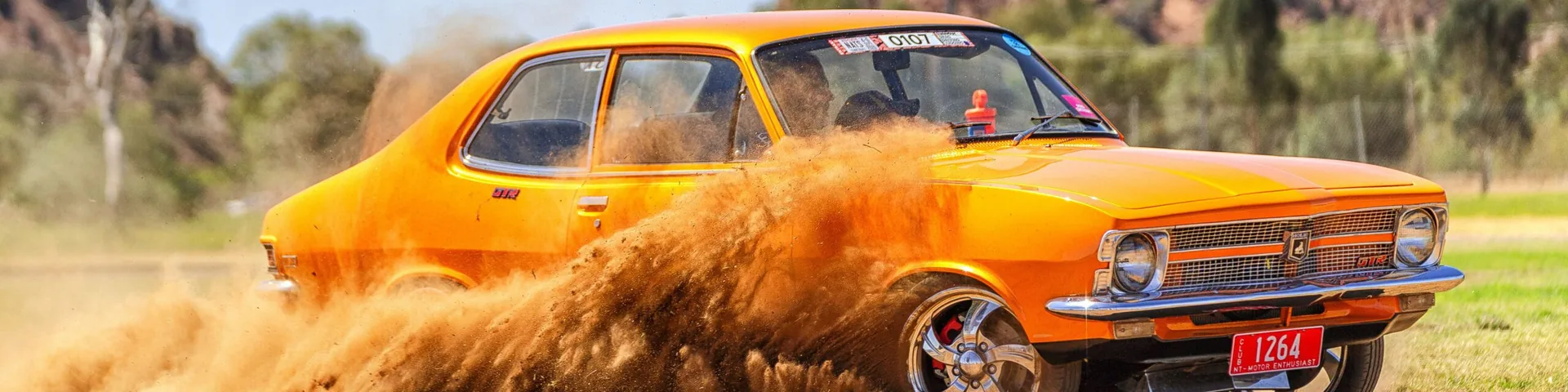 Orange sedan drifting sideways in a dirt drag, throwing up a large dust cloud, plate '1264' and entry number '0107' on the windscreen.