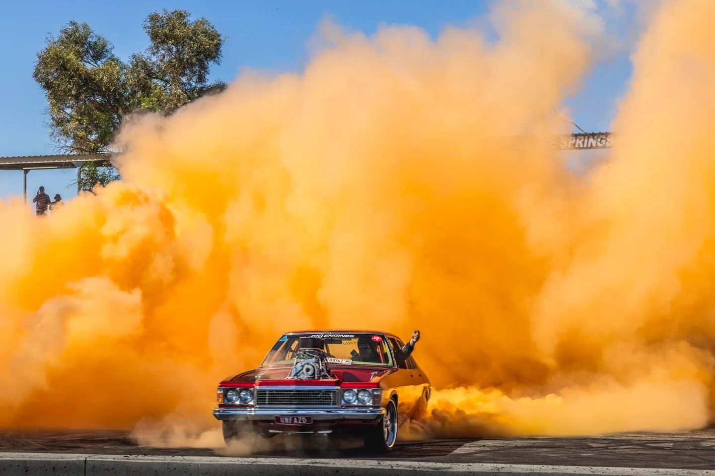 A dark red sedan, plate 'UNFAZD', vanishes into a thick orange burnout smoke cloud beside an Alice Springs sign, spectators watching from a small stand.